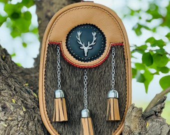 May include: A brown leather sporran with a black and silver stag head emblem. The sporran has three brown leather tassels attached to silver chains.