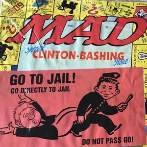 May include: A vintage MAD Magazine cover with the title "Clinton-Bashing Issue" in red. A pink insert reads "GO TO JAIL!" with a cartoon of Alfred E. Neuman. The background features a Monopoly-themed design.