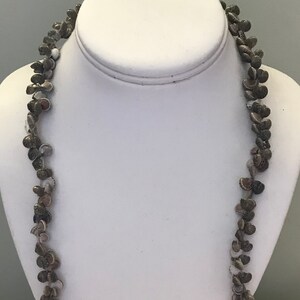 May include: A long necklace made of small, brown and white seashells. The necklace is strung on a thin cord and is displayed on a white mannequin.