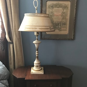 May include: A white and gold table lamp with a white lampshade featuring a gold leaf design. The lamp is on a dark wood nightstand with three drawers.