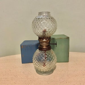 May include: A clear glass oil lamp with a textured, bubble-like pattern. The lamp has a round base and a spherical top, connected by a brass-colored metal piece. The lamp is sitting on a light pink surface.