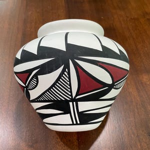May include: A white ceramic vase with a rounded body and a wide, slightly flared rim. The vase features a geometric design in black and red against a white background. The design includes triangles, lines, and curved shapes.