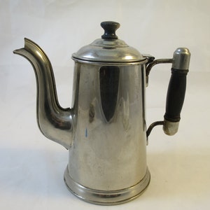 May include: A silver metal coffee pot with a black handle. The pot has a rounded body and a spout on the side.