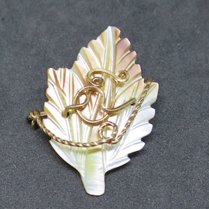 Peut inclure: Une broche en forme de feuille dorée avec un motif en fil doré en forme de lettre "L". La broche est en nacre et présente un jeu de couleurs blanc et or.