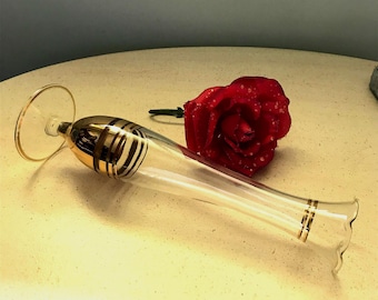 May include: A clear glass bud vase with a flared rim and a round base, featuring gold-colored horizontal bands around the neck and near the opening. A single red rose with visible water droplets on its petals rests beside the glass vase.