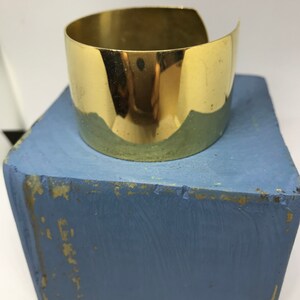 May include: A shiny gold metal cuff bracelet. The bracelet is wide and has a simple, rounded design.