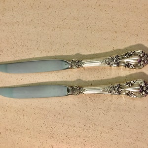 May include: Two silver butter knives with ornate handles. The knives have a polished finish and are in excellent condition.