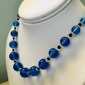 May include: A blue beaded necklace with large, round beads. The necklace features clear beads and small black accents between the larger blue beads. The necklace is displayed on a white jewellery bust.