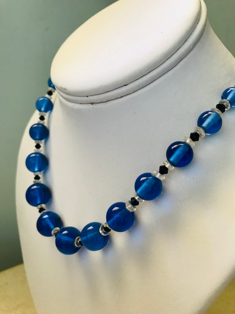 May include: A blue beaded necklace with large, round beads. The necklace features clear beads and small black accents between the larger blue beads. The necklace is displayed on a white jewellery bust.