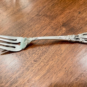 May include: Silverware fork with three tines and a decorative handle. The handle features floral and scrollwork designs. The fork is made of a shiny, reflective metal, likely silver or a silver-colored alloy. The fork is lying on a wooden surface.