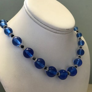May include: A blue beaded necklace with large, round beads and smaller accent beads. The necklace is displayed on a white jewellery bust. The beads are a vibrant blue colour, with black and clear accent beads.