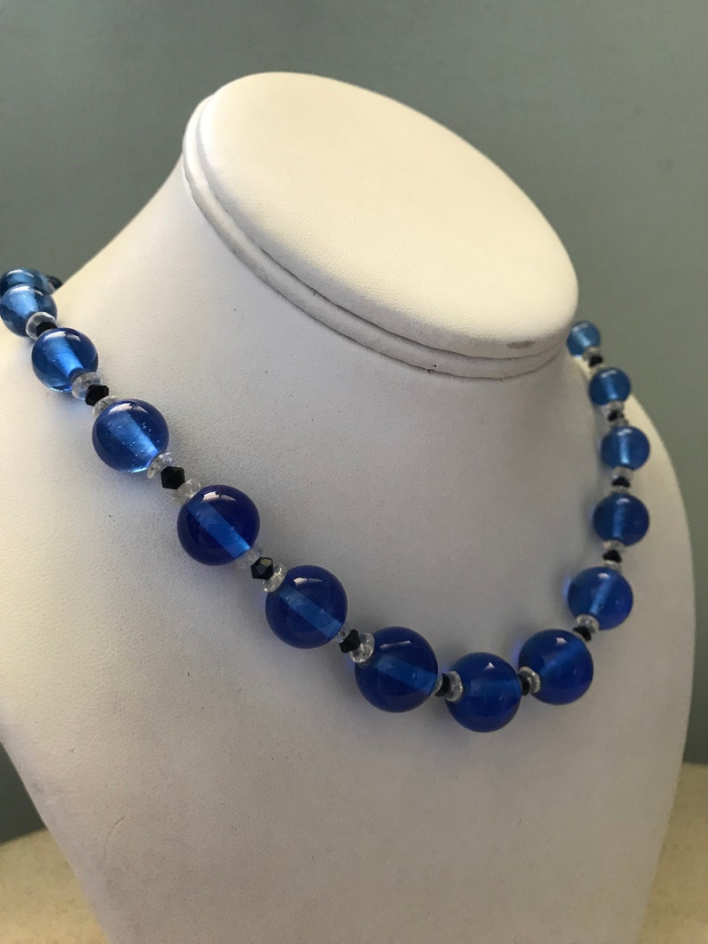 May include: A blue beaded necklace with large, round beads and smaller accent beads. The necklace is displayed on a white jewellery bust. The beads are a vibrant blue colour, with black and clear accent beads.