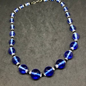 May include: A necklace with large, round, cobalt blue glass beads, with smaller clear and black beads. The necklace is strung on a black cord and has a silver-tone clasp. The beads have a translucent quality.