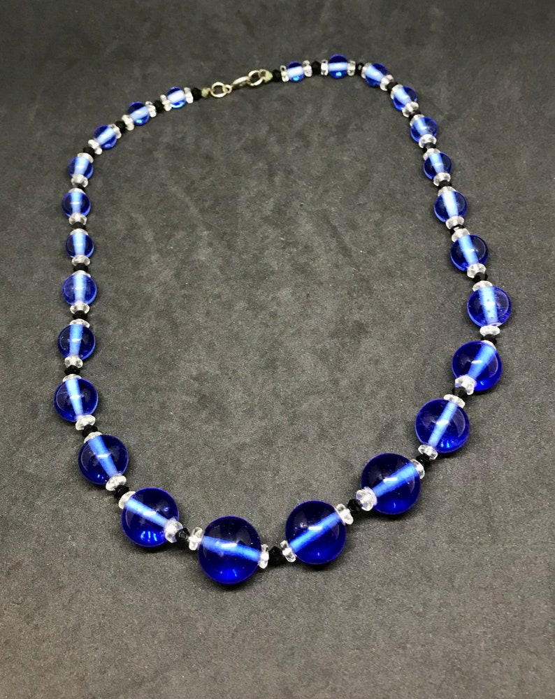May include: A necklace with large, round, cobalt blue glass beads, with smaller clear and black beads. The necklace is strung on a black cord and has a silver-tone clasp. The beads have a translucent quality.