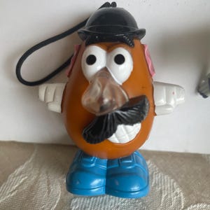 May include: A Mr. Potato Head toy figurine with a black hat, mustache, and blue shoes. The toy has white arms, pink ears, and a clear nose. A black string is attached to the top of the hat.
