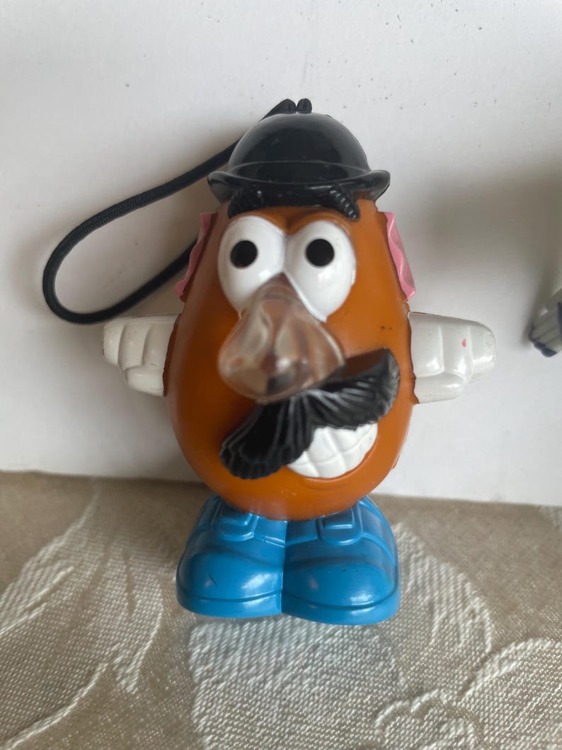 May include: A Mr. Potato Head toy figurine with a black hat, mustache, and blue shoes. The toy has white arms, pink ears, and a clear nose. A black string is attached to the top of the hat.
