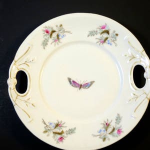 Puede incluir: Un plato ovalado blanco para servir con ribetes dorados y dise&ntilde;os florales. El plato tiene dos asas y una mariposa pintada en el centro.