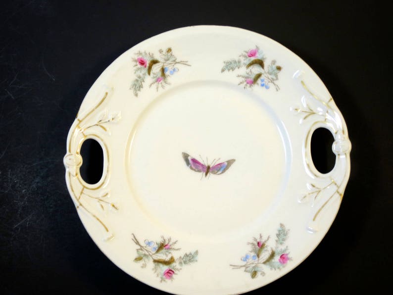 Puede incluir: Un plato ovalado blanco para servir con ribetes dorados y dise&ntilde;os florales. El plato tiene dos asas y una mariposa pintada en el centro.