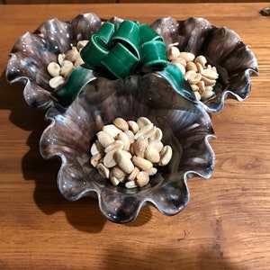 May include: A three-tiered ceramic serving dish with a brown and white glaze. The dish is decorated with a green bow and filled with peanuts.