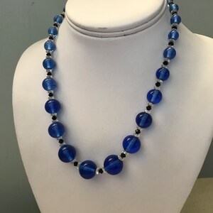 May include: A blue beaded necklace featuring large, round, cobalt blue beads and smaller black and clear beads. The necklace is displayed on a white mannequin bust.
