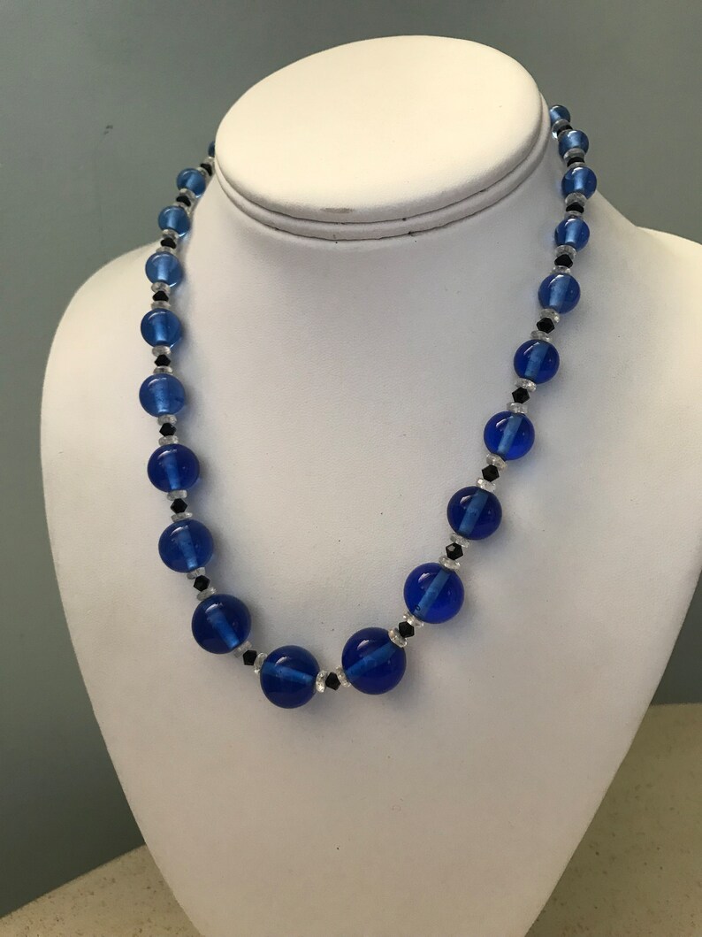 May include: A blue beaded necklace featuring large, round, cobalt blue beads and smaller black and clear beads. The necklace is displayed on a white mannequin bust.
