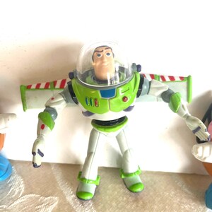 May include: A Buzz Lightyear action figure with a clear helmet, green and white spacesuit, and red, white, and green wings. The figure is flanked by two Mr. Potato Head toys, one on each side.