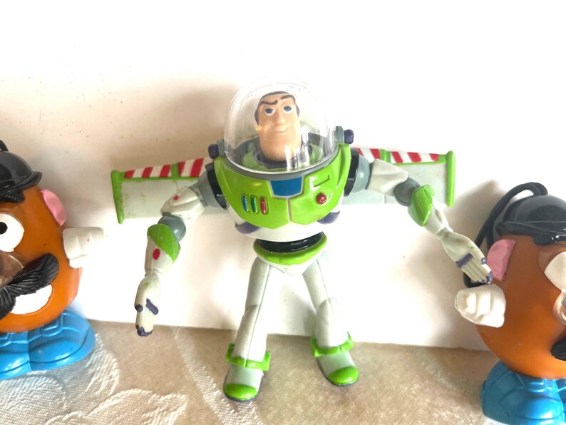 May include: A Buzz Lightyear action figure with a clear helmet, green and white spacesuit, and red, white, and green wings. The figure is flanked by two Mr. Potato Head toys, one on each side.