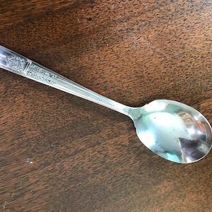 May include: A silver spoon with an ornate design on the handle. The spoon is on a wooden surface.