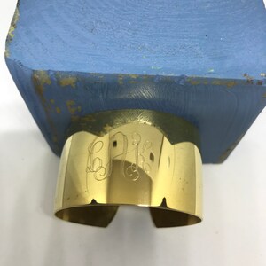 May include: A gold-colored metal cuff bracelet with an engraved monogram. The bracelet is sitting on a blue wooden surface.