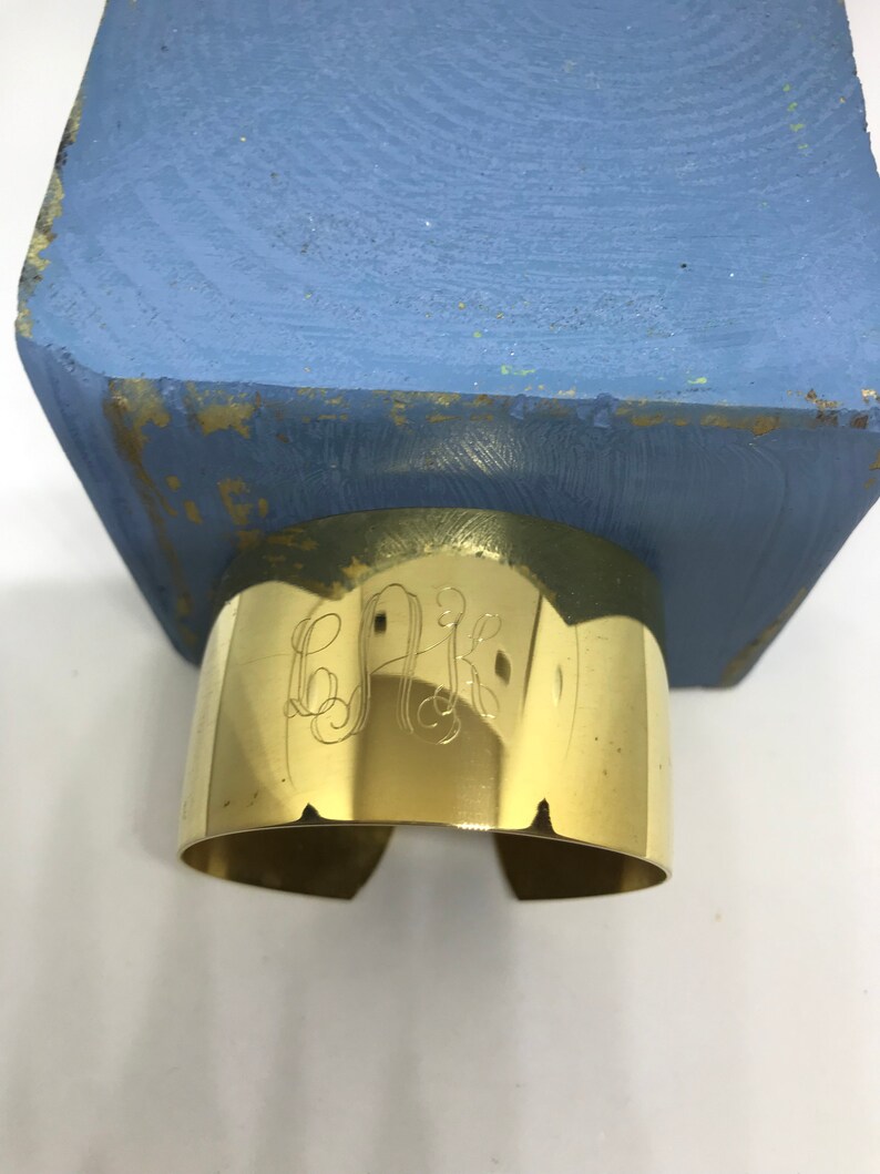 May include: A gold-colored metal cuff bracelet with an engraved monogram. The bracelet is sitting on a blue wooden surface.