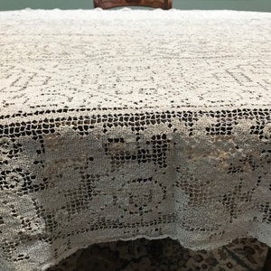 May include: A white crocheted tablecloth with a delicate floral pattern. The tablecloth is laid out on a wooden table.