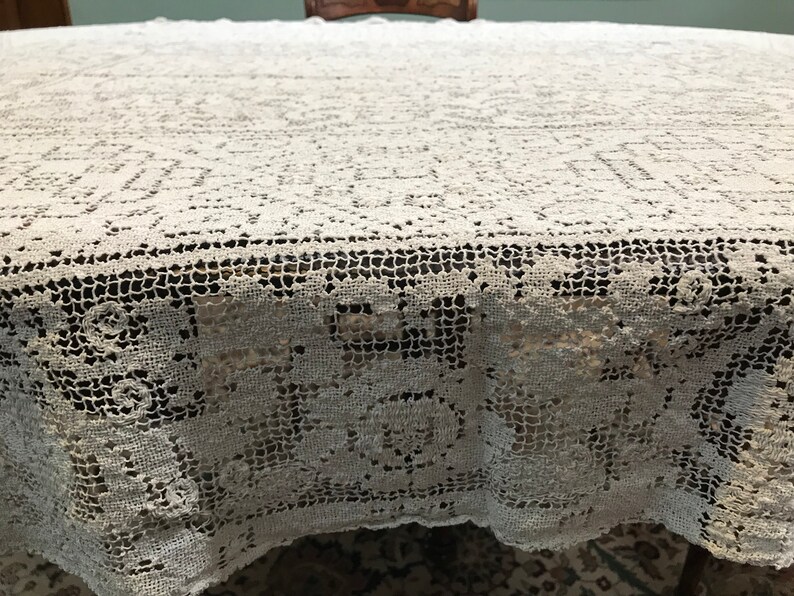 May include: A white crocheted tablecloth with a delicate floral pattern. The tablecloth is laid out on a wooden table.