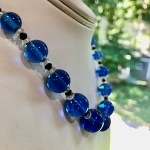 May include: A necklace featuring large, round, transparent blue beads. The design includes smaller clear and black beads interspersed between the larger blue beads. The necklace is displayed on a white mannequin.
