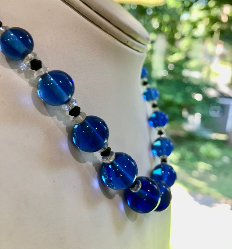 May include: A necklace featuring large, round, transparent blue beads. The design includes smaller clear and black beads interspersed between the larger blue beads. The necklace is displayed on a white mannequin.