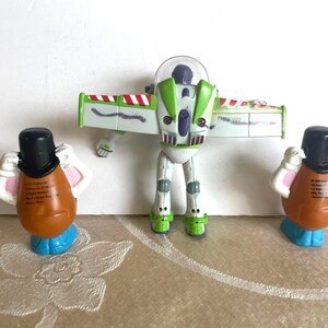 Pu&ograve; includere: Un gruppo di figurine di Toy Story. Buzz Lightyear, bianco e verde, con le ali spiegate. Due figure di Mr. Potato Head, marroni con cappelli neri e basi blu, affiancano Buzz. I giocattoli sono disposti su una superficie a motivi.