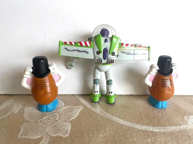Pu&ograve; includere: Un gruppo di figurine di Toy Story. Buzz Lightyear, bianco e verde, con le ali spiegate. Due figure di Mr. Potato Head, marroni con cappelli neri e basi blu, affiancano Buzz. I giocattoli sono disposti su una superficie a motivi.