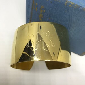 May include: A gold-toned metal cuff bracelet with an engraved monogram. The monogram is in a script font and features the letters "L.A.K."
