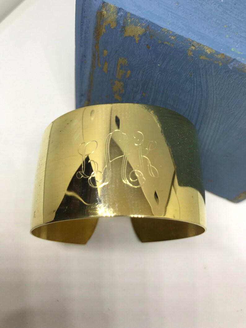 May include: A gold-toned metal cuff bracelet with an engraved monogram. The monogram is in a script font and features the letters "L.A.K."