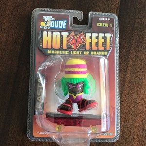 May include: A Tech Deck Hot Feet magnetic light-up fingerboard with a green and yellow Rastafarian-themed character on a red skateboard. The packaging features the text "Hot Feet" and "Magnetic Light-Up Boards".