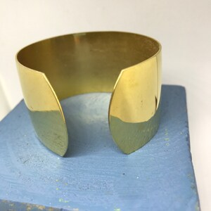 May include: A gold-colored metal cuff bracelet with a simple, open design. The bracelet is made of a single piece of metal that is bent into a circle and then has two small, pointed ends that are left open.