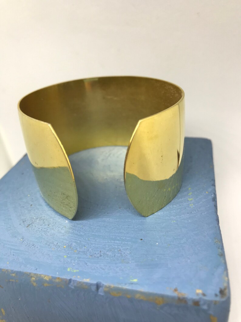 May include: A gold-colored metal cuff bracelet with a simple, open design. The bracelet is made of a single piece of metal that is bent into a circle and then has two small, pointed ends that are left open.