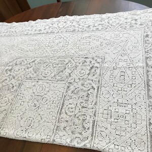 May include: White crocheted tablecloth with a delicate floral pattern. The tablecloth has a square shape and a lace border.