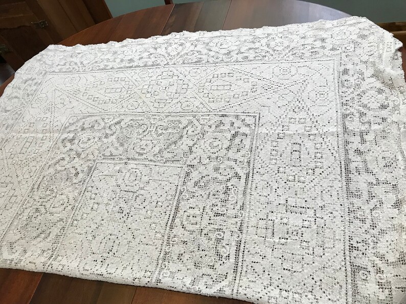 May include: White crocheted tablecloth with a delicate floral pattern. The tablecloth has a square shape and a lace border.
