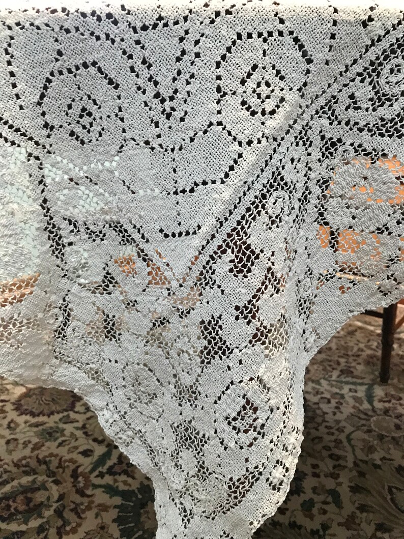 May include: White crocheted tablecloth with a delicate floral pattern. The tablecloth is made of fine lace and has a scalloped edge.