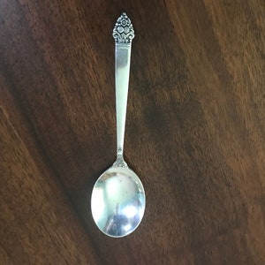 May include: A silver spoon with a floral design on the handle. The spoon has a rounded bowl and a long, slender handle. The handle is decorated with a detailed floral pattern at the top.
