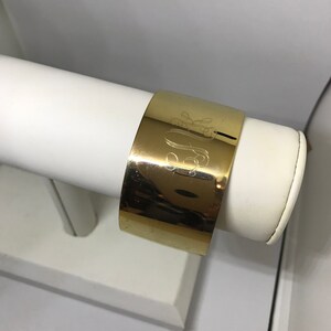 May include: A gold-toned metal cuff bracelet with an engraved monogram. The bracelet is displayed on a white stand.