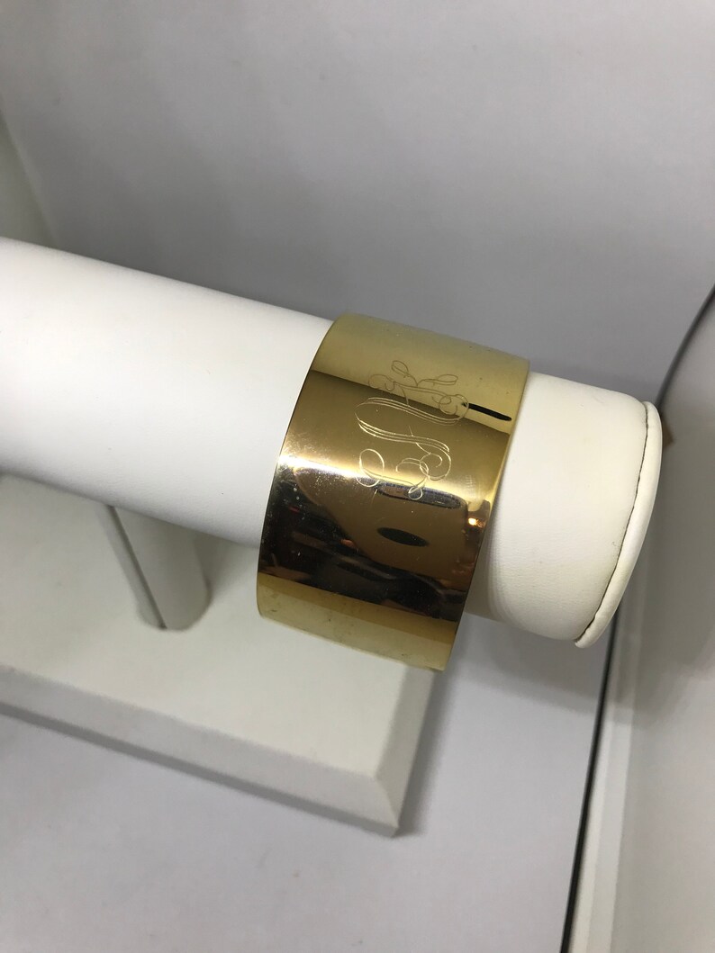 May include: A gold-toned metal cuff bracelet with an engraved monogram. The bracelet is displayed on a white stand.