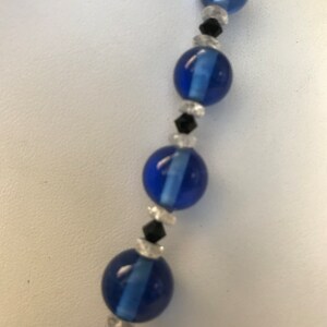 May include: A close-up of a necklace featuring large, round, cobalt blue beads interspersed with smaller black and clear beads. The necklace is displayed against a white background, highlighting the vibrant blue colour and intricate design of the jewellery.