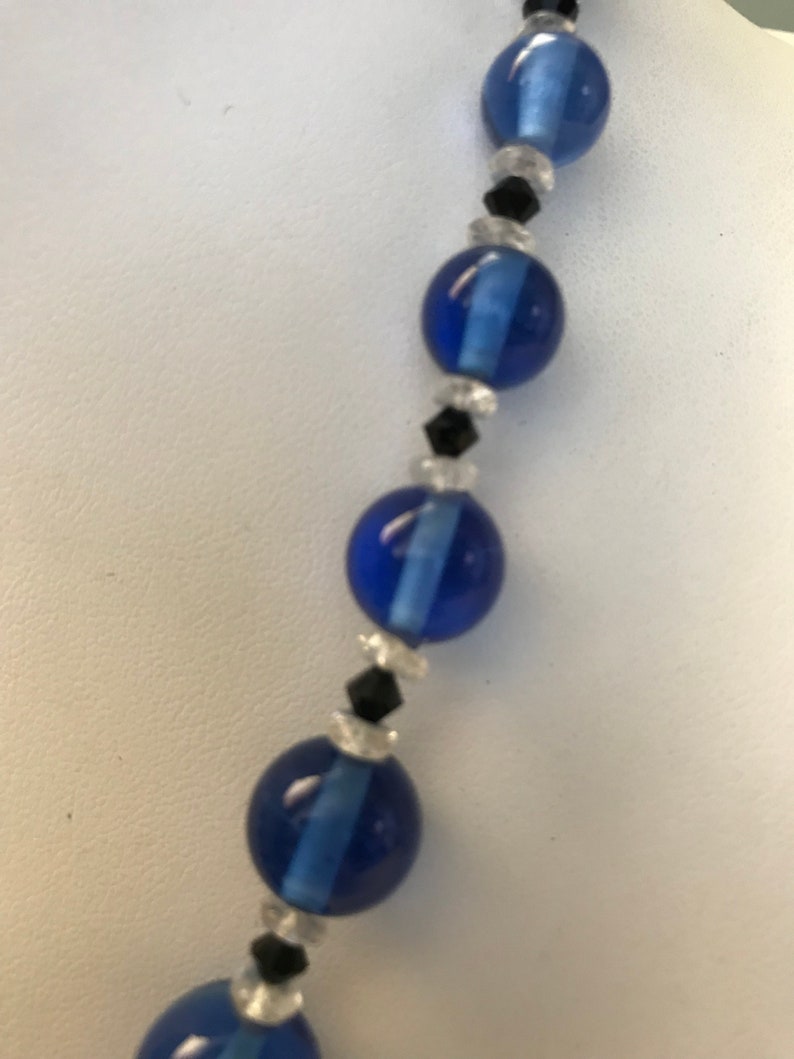 May include: A close-up of a necklace featuring large, round, cobalt blue beads interspersed with smaller black and clear beads. The necklace is displayed against a white background, highlighting the vibrant blue colour and intricate design of the jewellery.