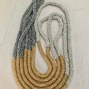 May include: A multi-strand beaded necklace with silver, gold, and white beads. The necklace is made of small, uniform beads and has a layered look.